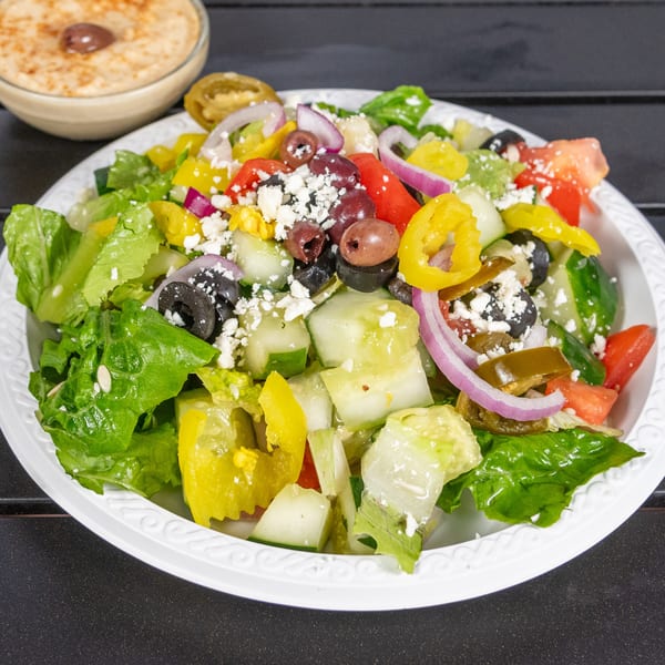 A vibrant Greek salad with lettuce, tomatoes, cucumbers, olives, feta, and peppers, with hummus in the background.