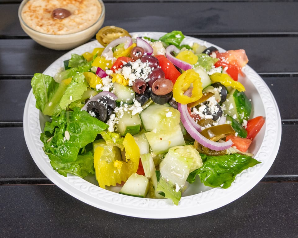 A vibrant Greek salad with lettuce, tomatoes, cucumbers, olives, feta, and peppers, with hummus in the background.