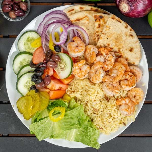 A vibrant shrimp platter with rice, pita bread, salad, red onions, and pickles on a dark table.