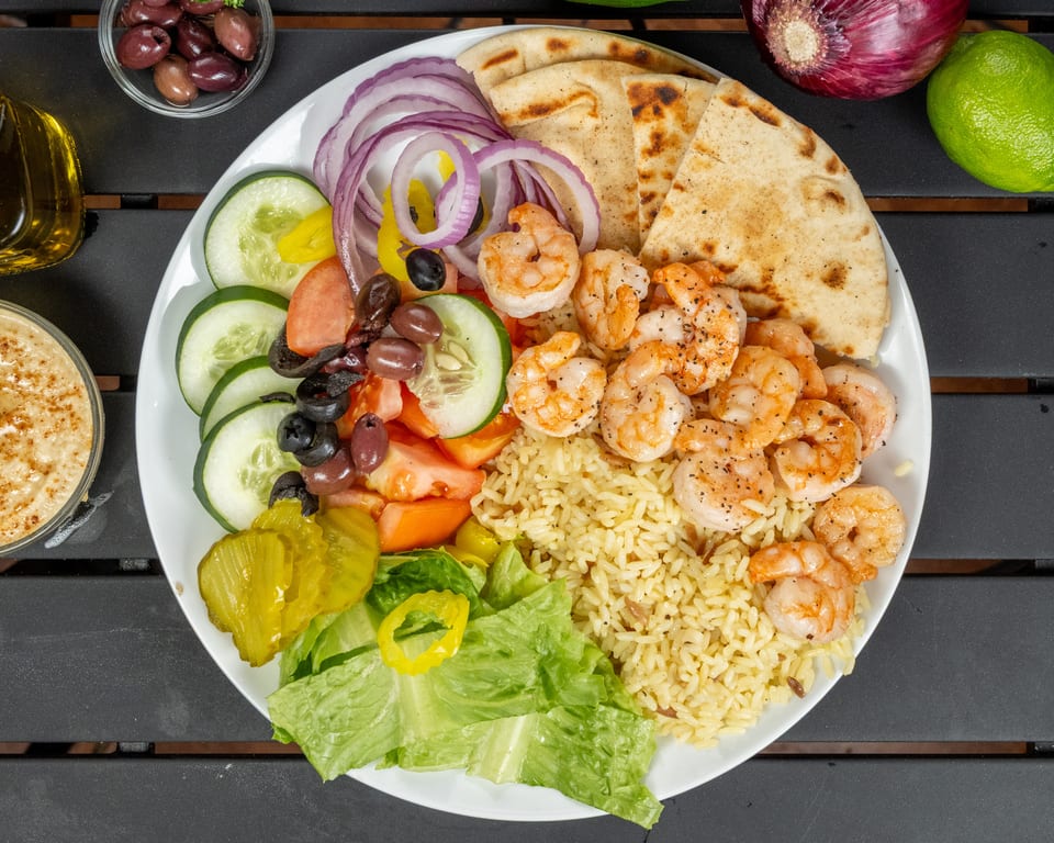 A vibrant shrimp platter with rice, pita bread, salad, red onions, and pickles on a dark table.