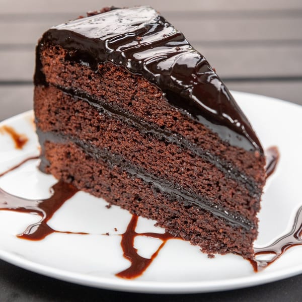 A rich slice of chocolate cake with dark chocolate ganache and syrup on a white plate.