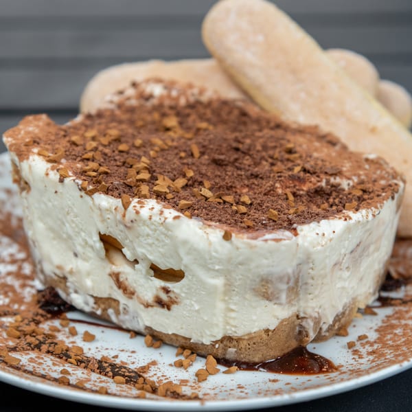A slice of tiramisu with ladyfingers, topped with cocoa powder and coffee crumbles on a white plate.