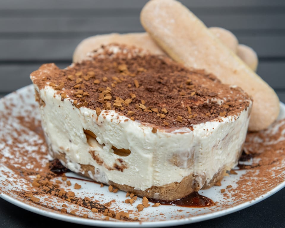 A slice of tiramisu with ladyfingers, topped with cocoa powder and coffee crumbles on a white plate.