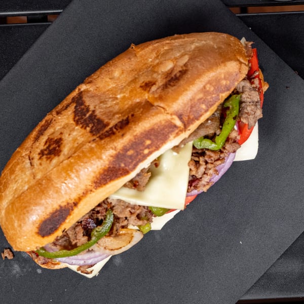 Overhead view of a cheesesteak sandwich with steak, cheese, peppers, and onions on a black tray.
