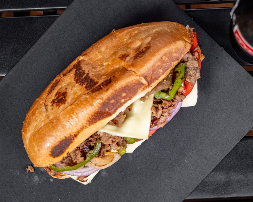 Overhead view of a cheesesteak sandwich with steak, cheese, peppers, and onions on a black tray.