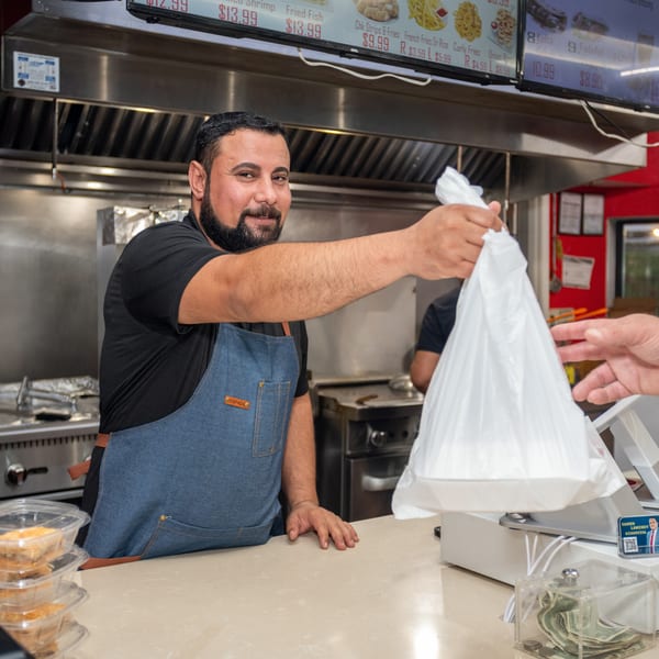 A smiling chef in an apron hands a customer a white takeout bag over a counter.