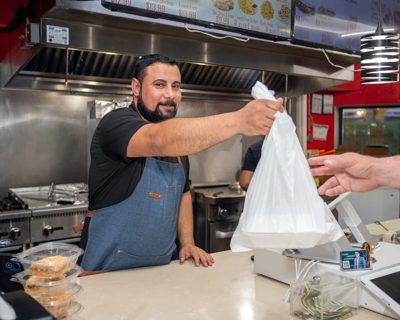 A smiling chef in an apron hands a customer a white takeout bag over a counter.