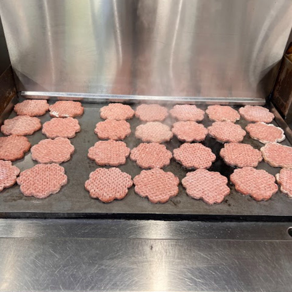 Rows of burger patties cooking on a large commercial griddle, with steam rising.