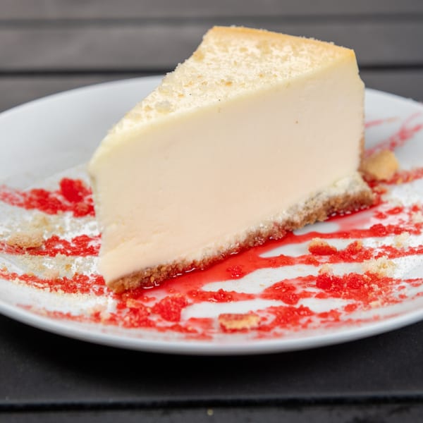 Slice of cheesecake with a graham cracker crust, drizzled with red berry sauce on a white plate.