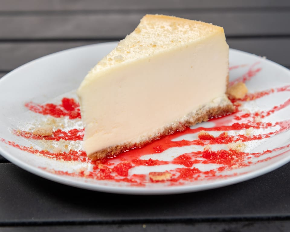 Slice of cheesecake with a graham cracker crust, drizzled with red berry sauce on a white plate.