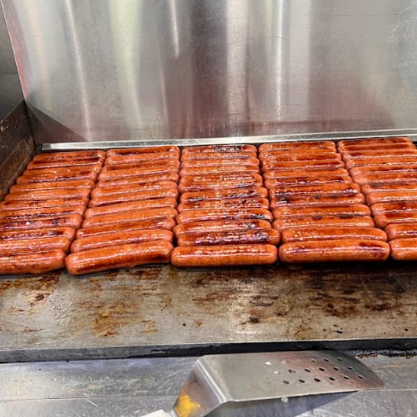 Many hot dogs cooking on a flat top griddle with a metal spatula nearby.