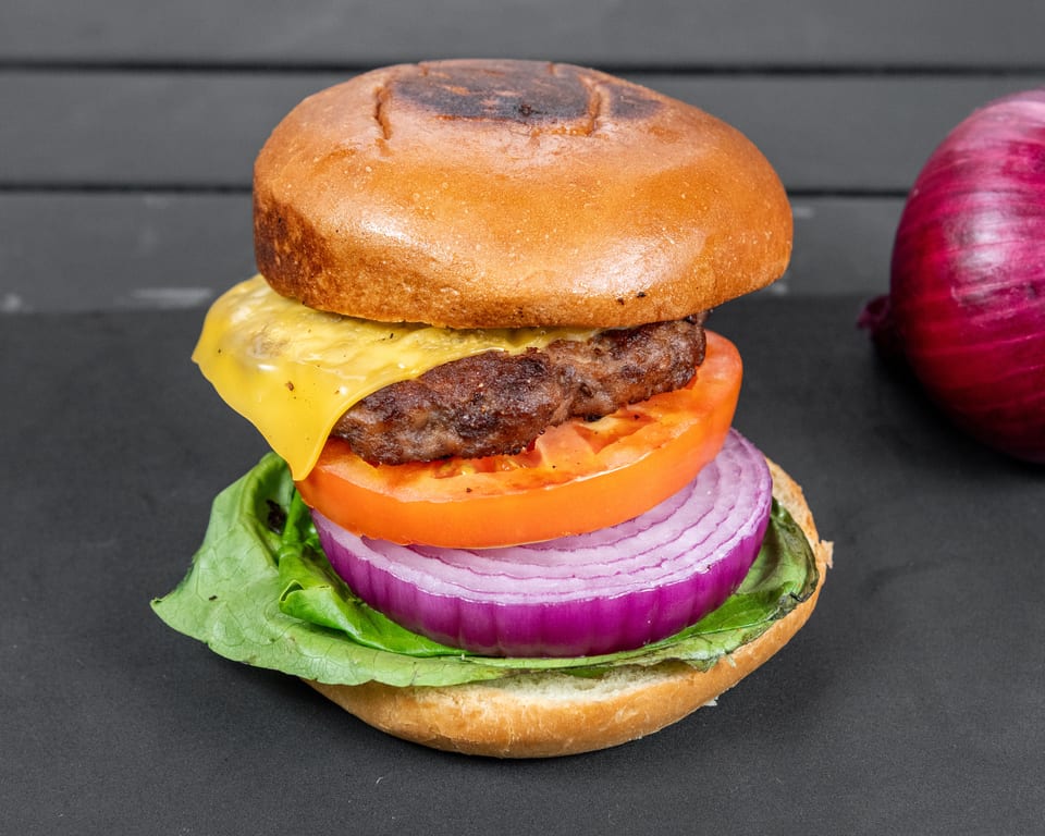 A cheeseburger with lettuce, tomato, and red onion on a dark surface, with a whole red onion beside it.