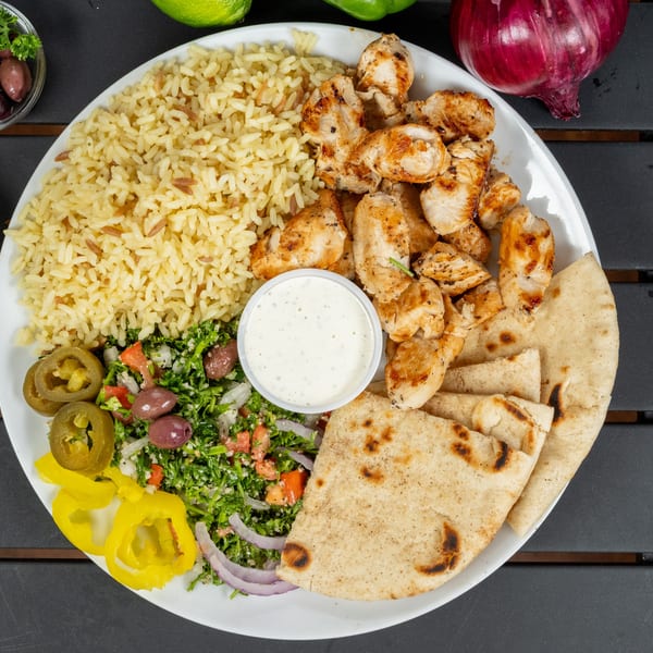 A white plate with grilled chicken, rice, tabbouleh salad, pita bread, and a side of sauce.