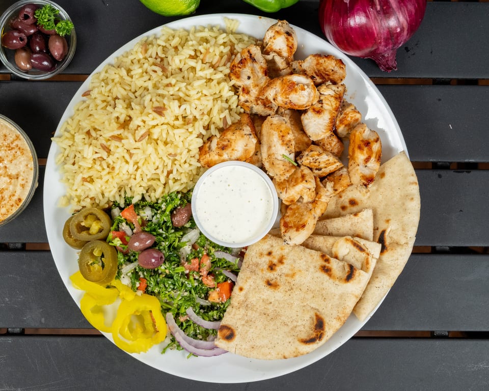 A white plate with grilled chicken, rice, tabbouleh salad, pita bread, and a side of sauce.