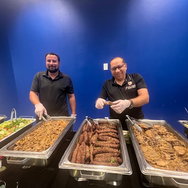 Two smiling Extra Dime Club Grill staff present a buffet of salads, rice, and grilled meats.