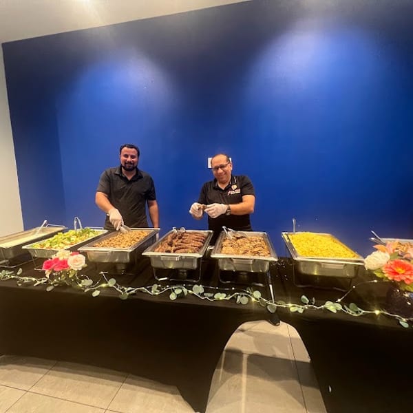 Two men serving food from a buffet line with various dishes and floral decorations.