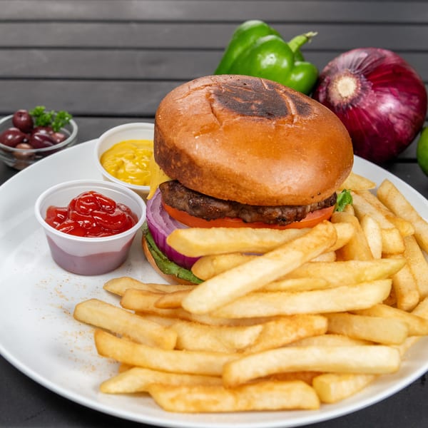 A burger with fries, ketchup, and mustard on a white plate, with vegetables in the background.