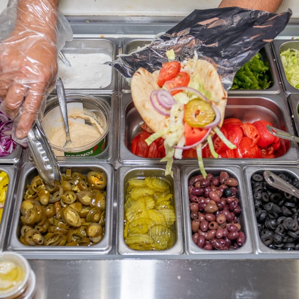 A person in gloves preparing a pita sandwich with various toppings from a buffet line.
