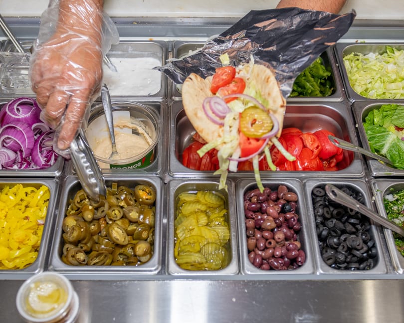 A person in gloves preparing a pita sandwich with various toppings from a buffet line.