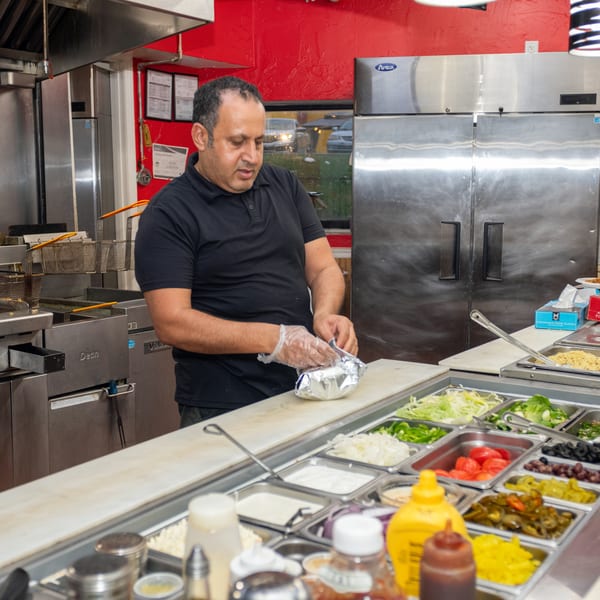A man in a black shirt wraps food in foil behind a counter with various fresh toppings.