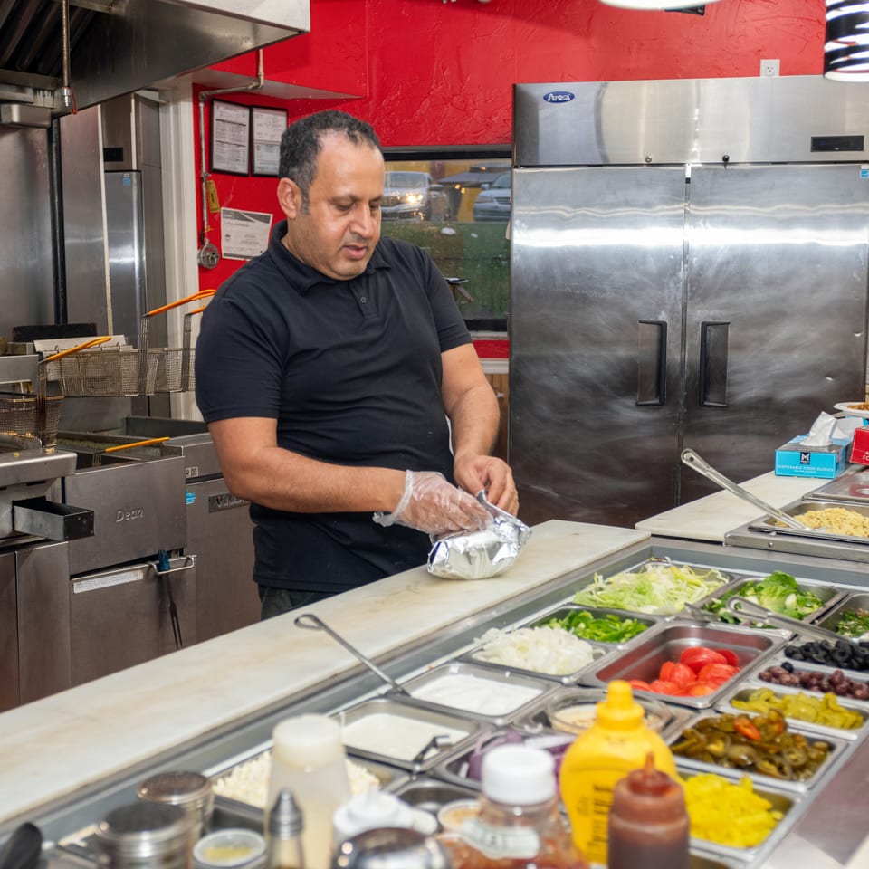 A man in a black shirt wraps food in foil behind a counter with various fresh toppings.