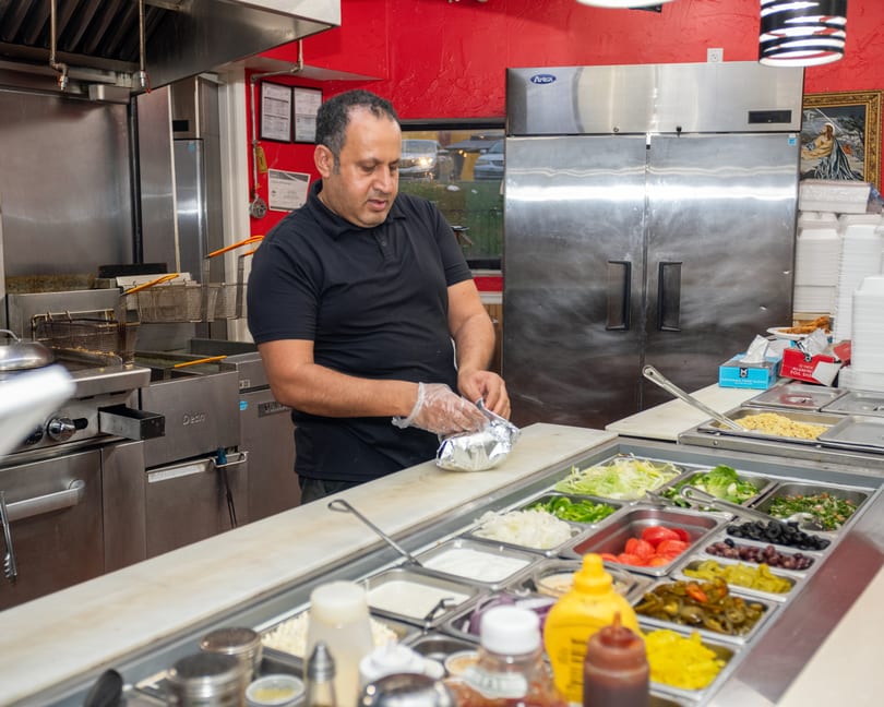 A man in a black shirt wraps food in foil behind a counter with various fresh toppings.