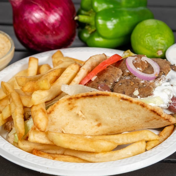 Gyro sandwich with fries on a white plate, with hummus and vegetables in the background.