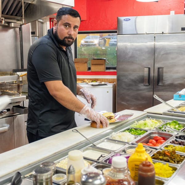 A chef in a commercial kitchen prepares a burger with fresh toppings at a counter.