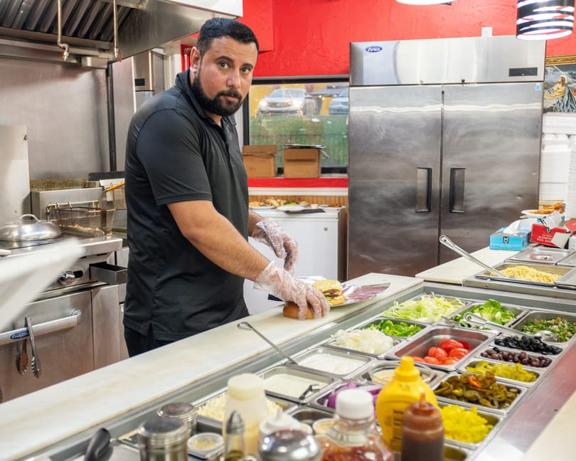A chef in a commercial kitchen prepares a burger with fresh toppings at a counter.