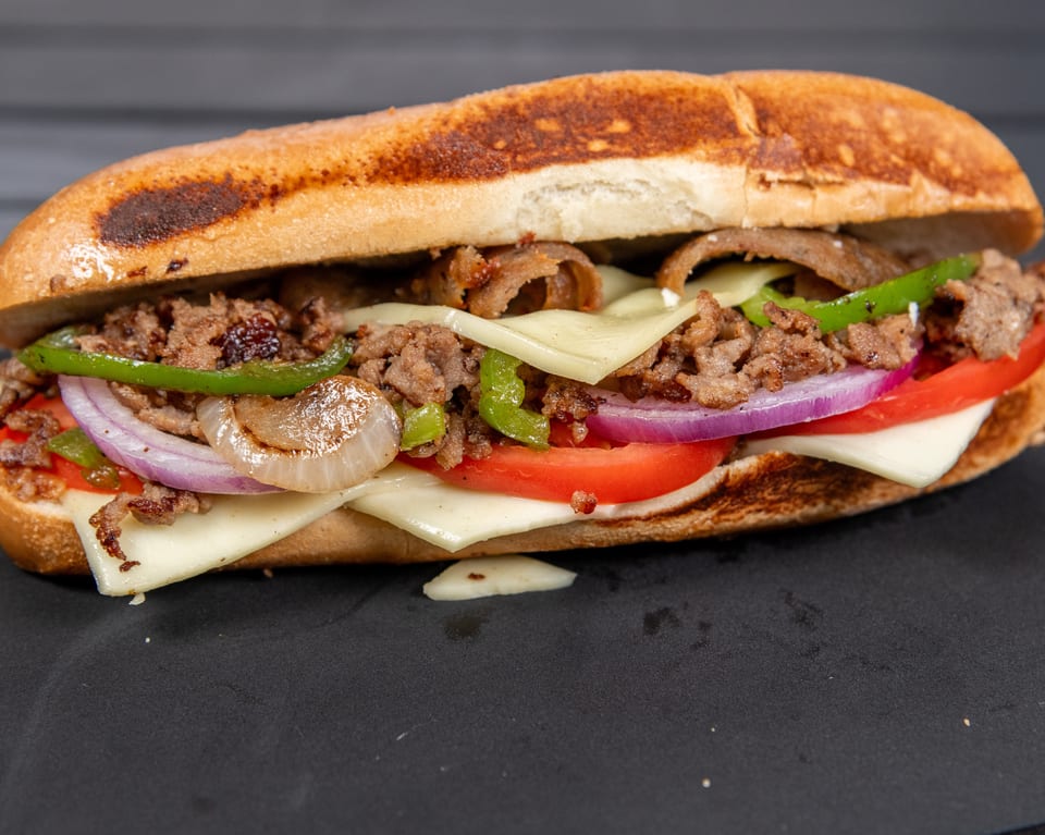 A close-up of a loaded cheesesteak sandwich with steak, cheese, peppers, onions, and tomatoes.